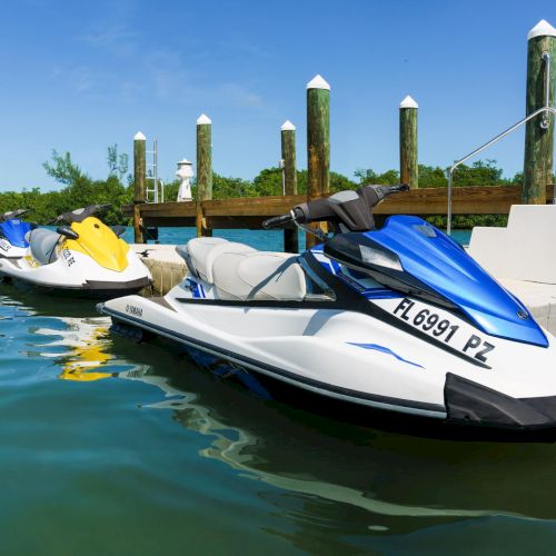 Jetskis docked at a wooden pier on calm blue water, bright sunny sky, tropical vibe. End.