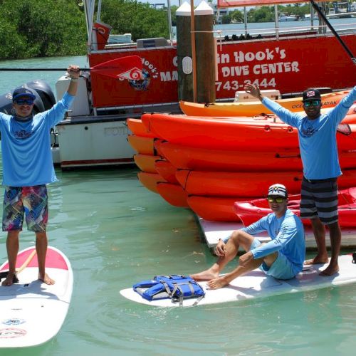 A swimmer stands on a dock next to two red boats, with a person in a blue shirt and another in blue shorts nearby, on a sunny day.