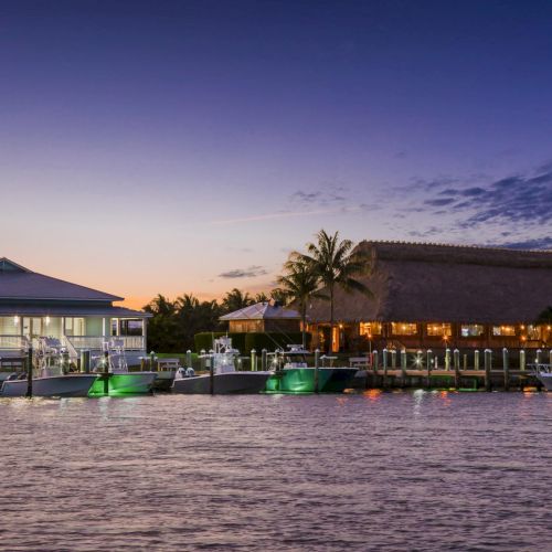 A picturesque waterfront at dusk with palm trees, two thatched-roof buildings on stilts, boats at a dock, and calm water reflecting the sunset, ending here.
