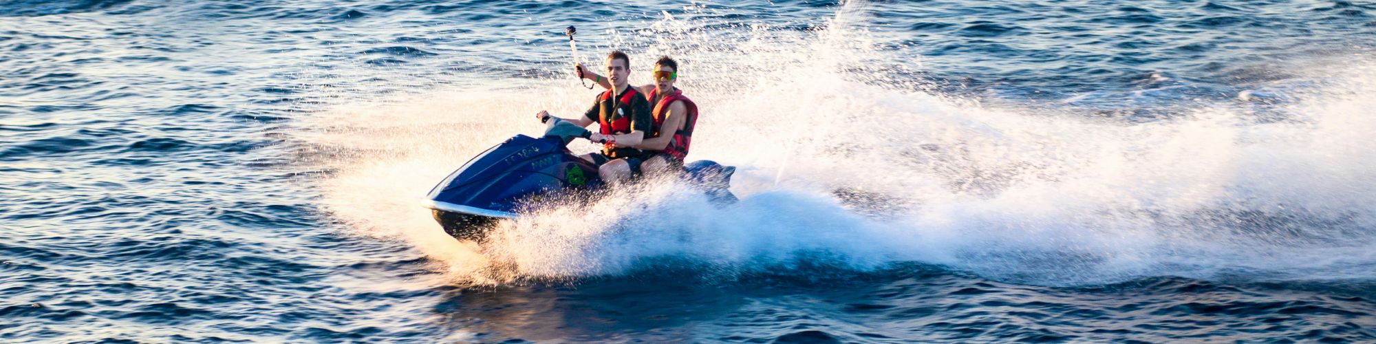 Two people ride a jet ski across choppy blue water as a second jet ski speeds nearby, spraying white foam and leaving a wake behind.